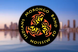 Federal judge dismisses Morongo tribe lawsuit challenging California gaming compact provisions. Morongo Band of Mission Indians circular logo with red and yellow tribal symbol on black background over blurred city skyline.