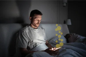 Man lying in bed at night looking at a glowing smartphone while stylized gold coin icons float upward from the screen, suggesting online gambling or betting activity as another person sleeps beside him in a dimly lit bedroom. Digital betting boom reshapes American gambling habits raising financial harm concerns